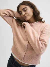 Νεαρή γυναίκα φορώντας μια μαλακή ροζ καμπαρντίνα με κουμπιά πάνω από ένα λευκό μπλουζάκι, πόζαρε με το ένα χέρι υψωμένο και το άλλο χέρι κοντά στο πρόσωπό της.