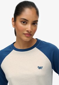 Beżowa koszulka z niebieskimi rękawami raglanowymi. Na piersi znajduje się małe niebieskie logo "Super Dry". Gładka tekstura materiału.