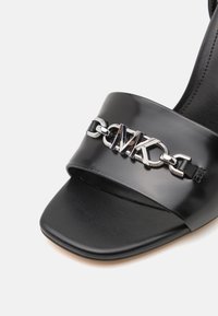 Black patent leather sandal with a wide strap featuring a silver logo chain accent. Square toe and smooth texture. Minimalistic design.