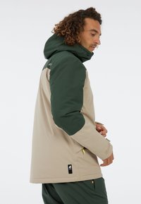 Béžová a tmavozelená vonkajšia bunda s kapucňou, ktorá má zipsové vrecká a textúrovanú tkaninu. Logo nášivka pri spodnom leme.
