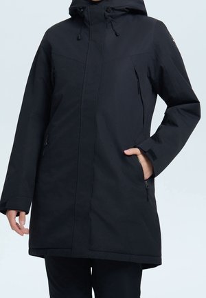 Persoon die een lange zwarte winterjas met capuchon en ritssluitingzakken draagt, hand in één zak, staand tegen een effen achtergrond.