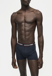 Homme mince à la peau foncée portant un boxer bleu marine Jack & Jones sur un fond clair uni, torse et parties supérieures des jambes visibles.