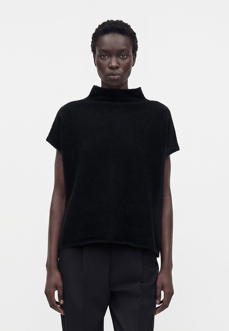 Femme aux cheveux courts portant un haut noir ample sans manches avec un col montant et un pantalon noir sur un fond blanc uni.