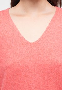 Πουλόβερ με V-λαιμόκοψη σε ανοιχτό κοραλί, μαλακή πλεκτή υφή, μικρές βελονιές, διακριτική γυαλάδα, με ακατέργαστη λεπτομέρεια στο τελείωμα του λαιμού.