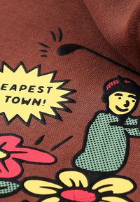 Smeđa tkanina s tiskanim grafikama; uključuje crtanog lika, cvjetne motive i oblačić za govor u kojem piše "NAJJEFTINIJI GRAD!" u žutoj boji.