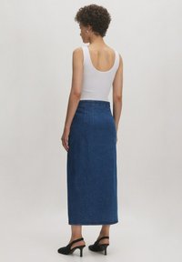 Denim midi-rok in donkerblauw met een aangesloten taille, zijzakken en tot halverwege de kuit. Gecombineerd met een wit tanktop en zwarte hakken.