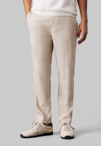 Pantaloni beige con una texture liscia, taglio dritto e tasche laterali, abbinati a sneakers chiare con suole scure.
