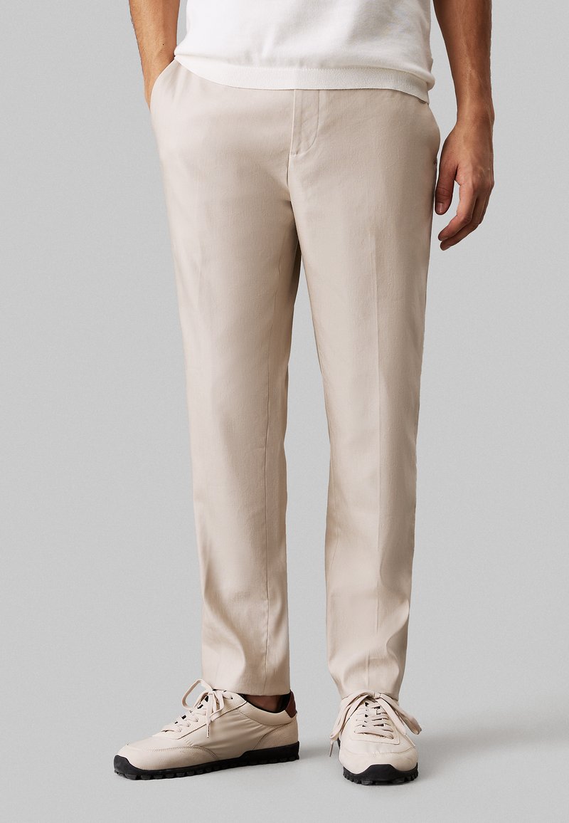 Pantaloni beige con una texture liscia, taglio dritto e tasche laterali, abbinati a sneakers chiare con suole scure.