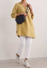 Gelbe Leinentunika mit Kragen, weiße Cigarette-Hosen und graue Umhängetasche mit gemustertem Riemen, kombiniert mit hellgrauen Sneakers.