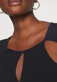 Svart topp med rund halsringning och ett nyckelhålssnitt, tillverkad av ett slätt tyg med en aning lyster. Guldörhängen i form av ringar kompletterar looken.