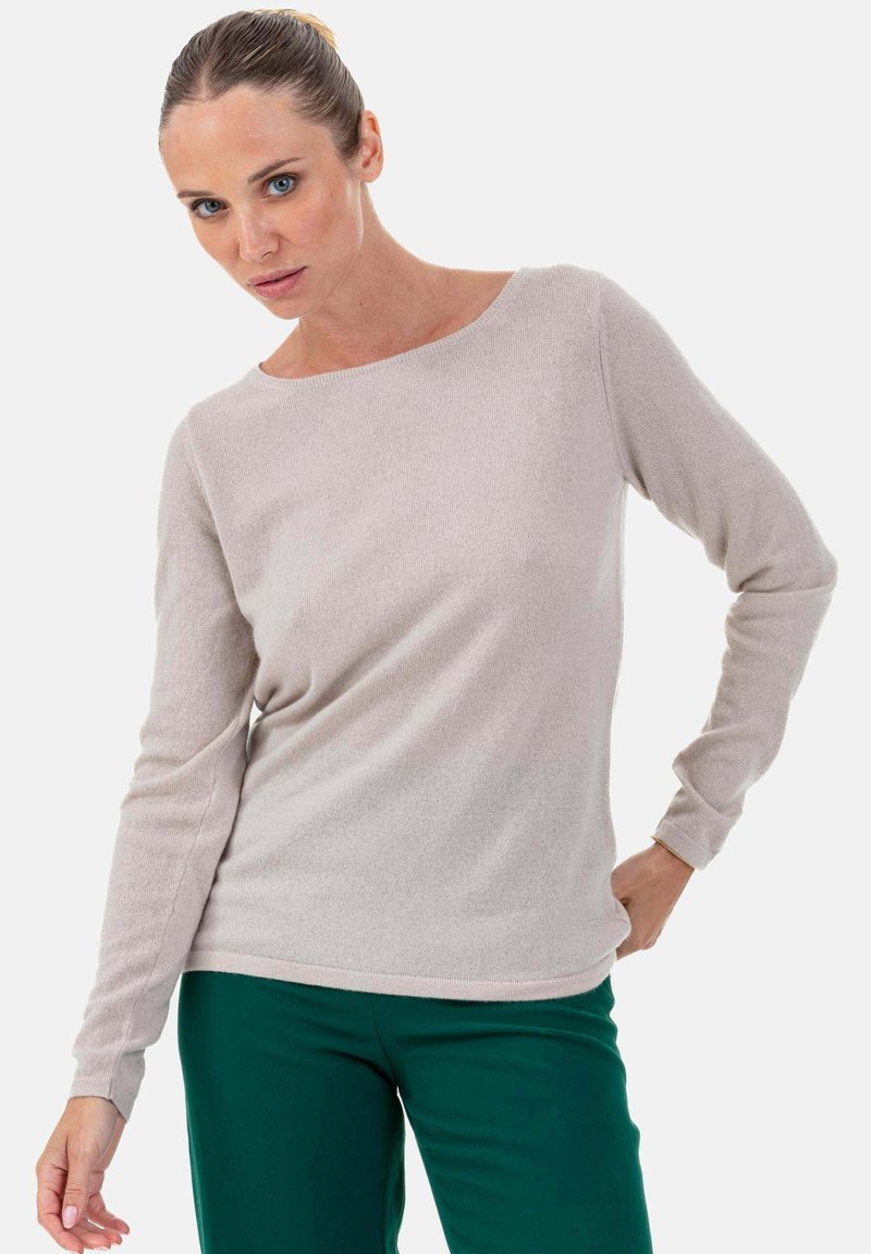 Femme aux cheveux attachés, portant un pull à manches longues beige clair et un pantalon vert foncé, posant avec une main sur la hanche.