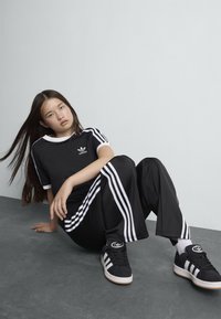 Survêtement noir Adidas avec des rayures blanches, haut à manches courtes et pantalon long, associé à des baskets noires avec des rayures supplémentaires et un logo.