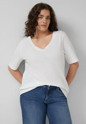 Kobieta o kręconych włosach, ubrana w biały top z krótkim rękawem i dekoltem w serek oraz niebieskie dżinsy, stoi z rękami za plecami na jednolitym szarym tle.