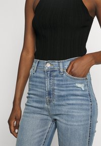 Top halter negro de canalé combinado con jeans de mezclilla azules de tiro alto que presentan un detalle desgastado y un cierre de botón clásico.