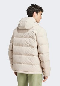 Beige puffer jacket with horizontal quilted sections, attached hood, and soft texture, worn over light green pants, shown from the back.