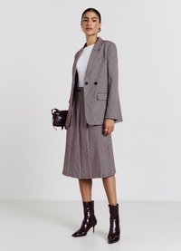 Blazer gris à motif pied-de-poule avec un design croisé, haut blanc, jupe midi plissée, et bottines bordeaux brillantes. Tient un petit sac.