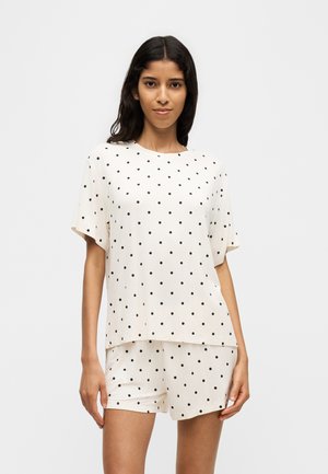 Vrouw draagt een witte top met korte mouwen en bijpassende korte broek met zwarte stippen, staand tegen een effen lichte achtergrond.