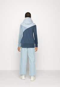Felpa con cappuccio in sfumature di blu, caratterizzata da un cappuccio superiore testurizzato e una sezione inferiore scura in contrasto. Abbinata a pantaloni a gamba larga blu chiaro.