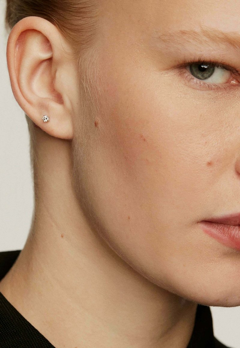 Pendiente de plata con una piedra preciosa clara, situado sobre un tono de piel claro. Primer plano de la oreja que muestra una textura mínima y color natural.