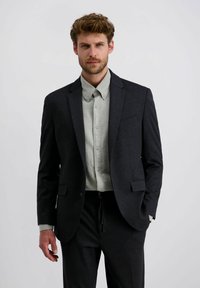 Man met krullend haar in een donker colbert over een lichtgrijze overhemd met knopen en bijpassende donkere broek tegen een egale achtergrond.