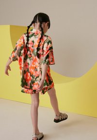 Cămașă cu imprimeu floral, cu mânecă scurtă, în nuanțe vibrante de portocaliu și verde, asortată cu pantaloni scurți. Sandale negre și gri completează ținuta.