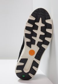 Svart sneaker med en texturerad svart ovandel och en vit gummisula med ett distinkt mönster och orange logotypdetalj.