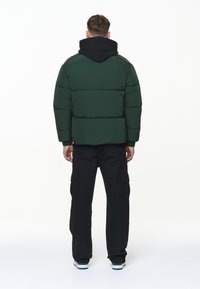 Chaqueta tipo puffa en verde oscuro con capucha negra, que presenta un diseño acolchado, puños acanalados y un corte relajado, combinada con pantalones cargo negros.