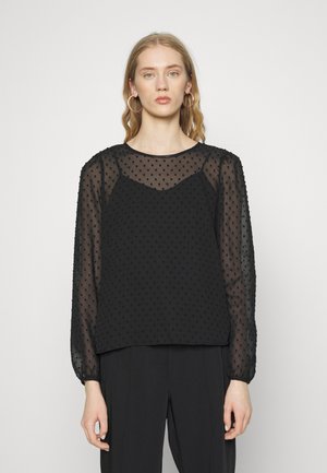 Blusa preta transparente com bolinhas texturizadas, mangas longas bufantes e decote redondo. Usada em camadas sobre uma blusa correspondente.