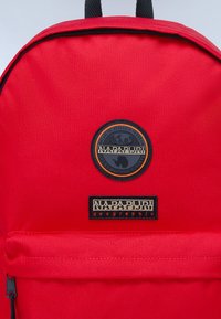 Zaino rosso realizzato in tessuto resistente, con una toppa logo circolare e un'etichetta di branding rettangolare in nero e oro sulla tasca frontale.