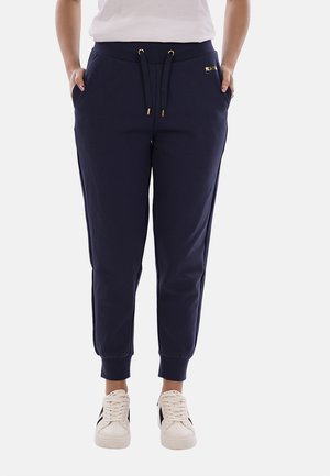 Donna che indossa pantaloni jogger blu navy con vita con coulisse e polsini elasticizzati, abbinati a sneakers bianche con strisce nere.