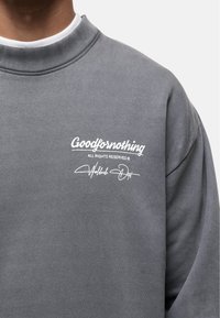Felpa grigia con colletto a coste. Presenta la scritta bianca "Goodfornothing" e la stampa distintiva sul davanti. Texture morbida, design casual.