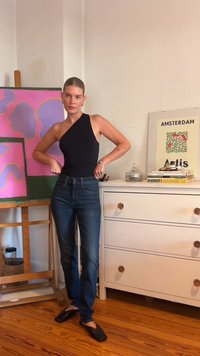 Μαύρο bodysuit με έναν ώμο, συνδυασμένο με σκούρο μπλε τζιν και μαύρα παπούτσια με τετράγωνη μύτη. Ξύλινο πάτωμα και πολύχρωμες ζωγραφιές στο φόντο.