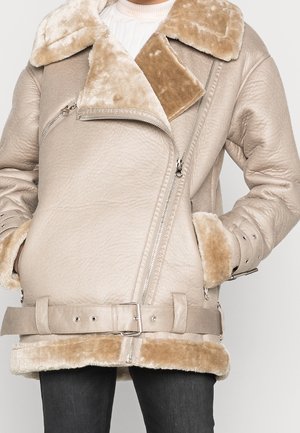 Beige fåreskindsjakke med bred blød krave, asymmetrisk lynlås, bælte i taljen og bløde manchetter, båret over en hvid sweater og sorte bukser.
