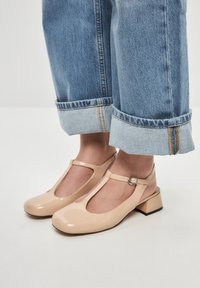 Beige kiiltoherkkä T-nauhashoes matalalla lohkokannan ja leikattu muotoilu, yhdistettyinä käännettyihin vaaleansinisiin farkkuihin.