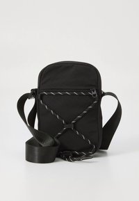 Melna auduma somiņa ar plecu siksnu, kurai ir rāvējslēdzējs augšpusē un baltas izšūšanas detaļas. Kompakts, taisnstūrveida formāts.