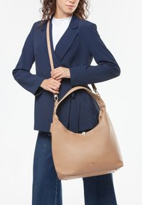 Beige leren handtas met een relaxte silhoeët, glanzend metalen onderdelen en een schouderband, gecombineerd met een marineblauwe blazer en denim.