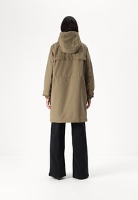 Khaki parka bunda s kapucí, elastickými manžetami a detailem na zadním díle, kombinovaná s černými širokými kalhotami. Hladká, odolná textilie.