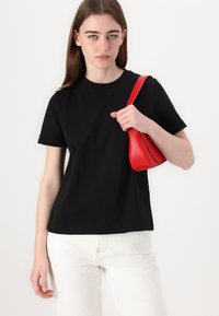 Svart kortermet T-skjorte i mykt stoff, kombinert med hvite jeans. Holder en liten rød skinnhåndveske med avrundet design og glatt overflate.