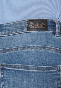 Vaqueros de mezclilla azul con un lavado claro, que presentan una etiqueta negra con letras doradas, diseño de cinco bolsillos y costuras en contraste.