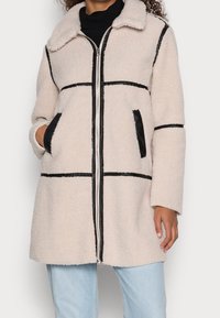 Chaqueta de forro polar en beige claro con ribetes negros, que presenta un cierre de cremallera en el frente, cuello y dos bolsillos laterales. Textura suave y corte oversized.