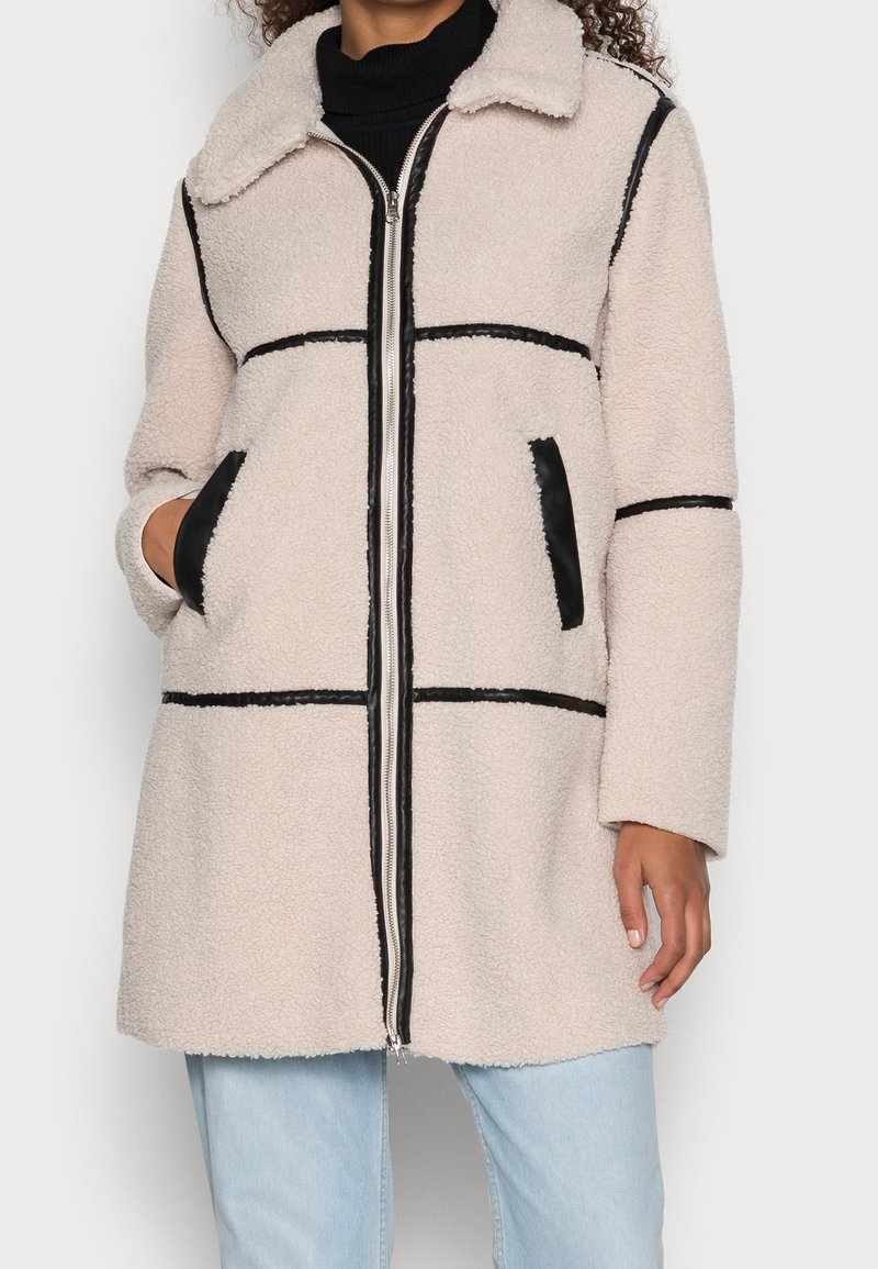 Chaqueta de forro polar en beige claro con ribetes negros, que presenta un cierre de cremallera en el frente, cuello y dos bolsillos laterales. Textura suave y corte oversized.