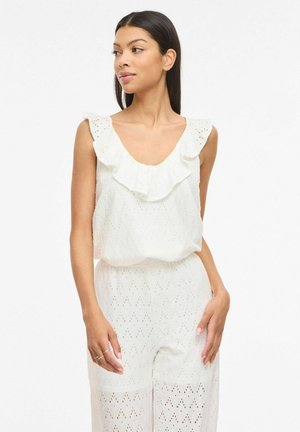 Femme portant un haut blanc sans manches en œillet avec un col à volants et un pantalon assorti, debout sur un fond blanc.