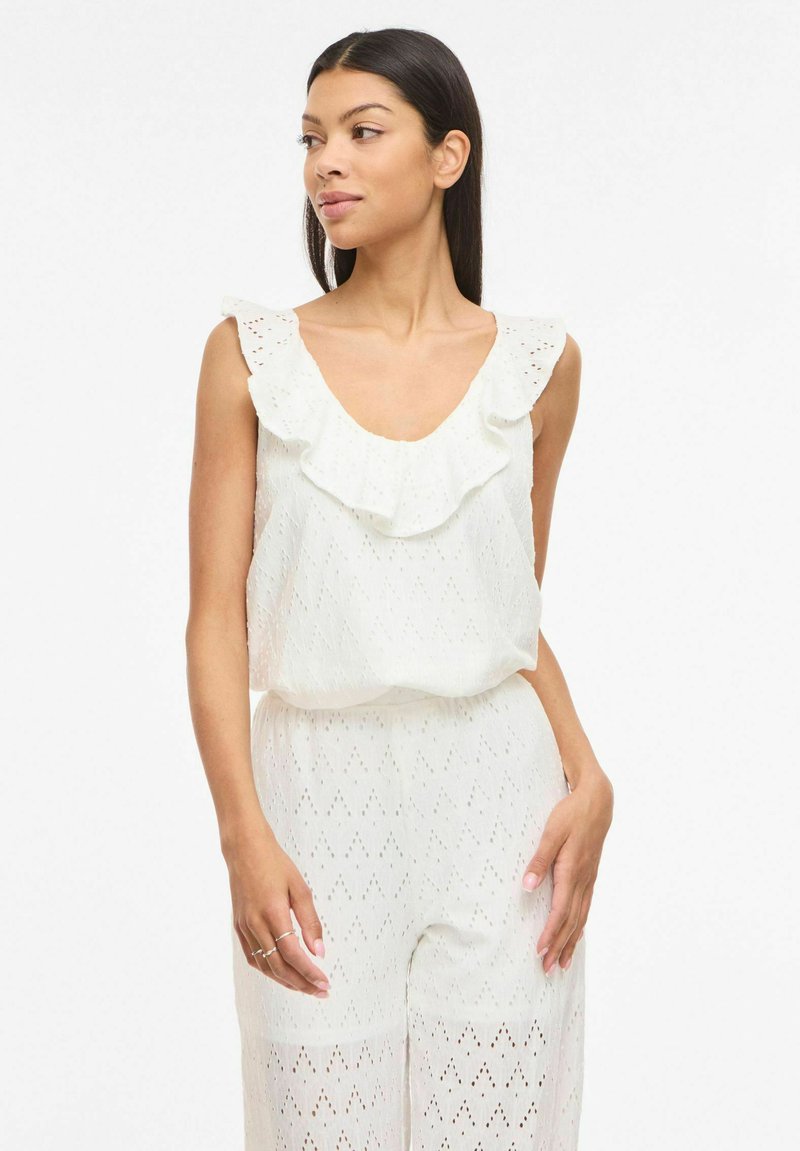 Femme portant un haut blanc sans manches en œillet avec un col à volants et un pantalon assorti, debout sur un fond blanc.