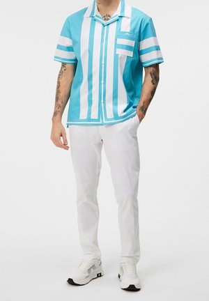 Mann trägt ein kurzärmeliges türkis-weiß gestreiftes Hemd mit weißen Hosen und weißen Turnschuhen, steht mit einer Hand in der Tasche.