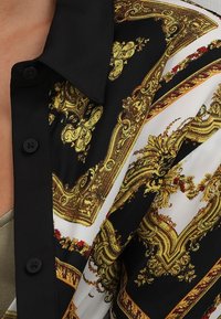 Černá košile s límečkem, zdobená zlatým barokním vzorem a knoflíky, vyrobená z lesklé látky. Design se střídá mezi černými a bílými částmi.