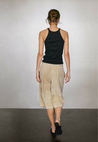Černý tílko, béžové široké kalhoty z měkké látky a černé nazouvací boty. Model stojí zády k neutrálnímu pozadí.