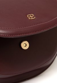 Burgundowa skórzana torebka w zaokrąglonym kształcie, z logo w kolorze złotym i zapięciem na zatrzask. Gładka tekstura z widocznymi detalami szycia.