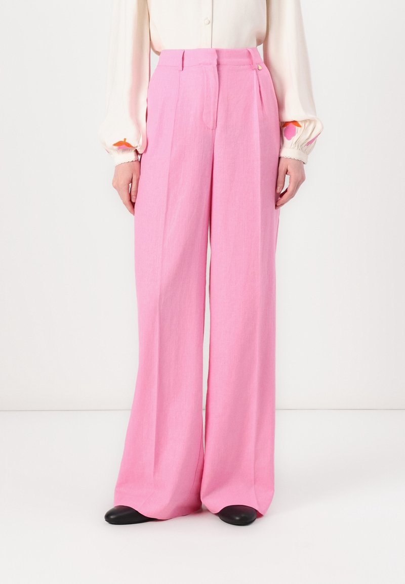 Personne portant un pantalon rose taille haute à jambes larges associé à un chemisier blanc à manches longues et des chaussures noires.