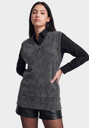 Kvinde iført en grå ærmeløs strikvest over en sort langærmet skjorte og sorte lædershorts, poserende med den ene hånd på hoften.