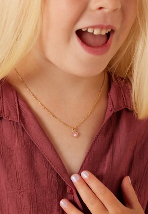 Gouden ketting met een roze hart hanger met een wit binnenhart ontwerp. Draag het met een gestructureerd bordeauxrood shirt.